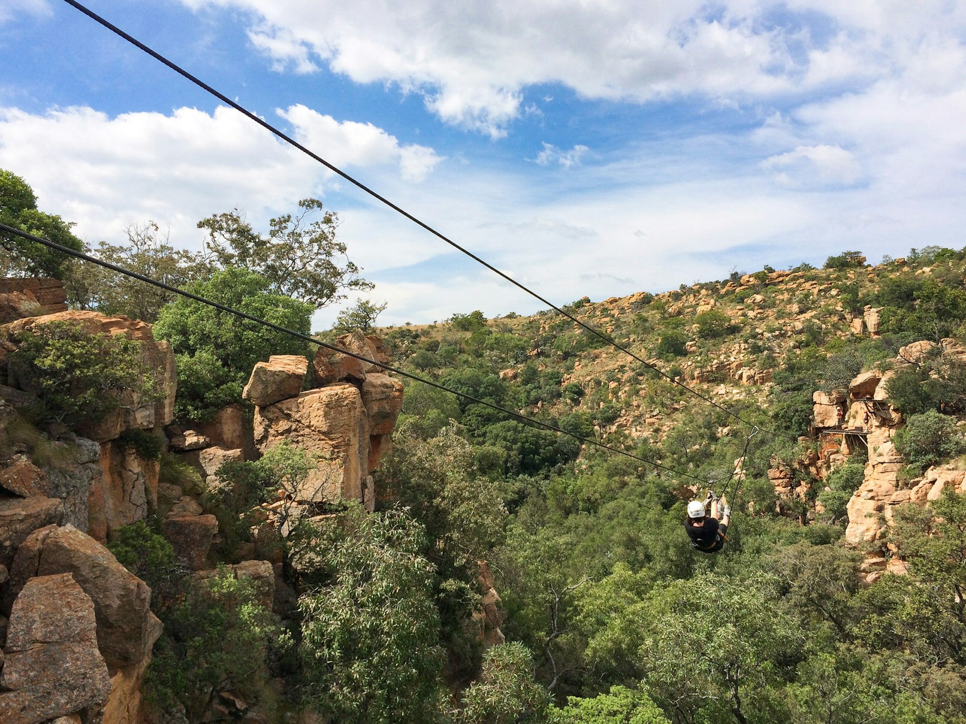 Two steel cables descend from the top left of the image to the bottom right; below them is a rocky and forested canyon along with a single person zooming downward on the cables as part of the zipline canopy tour
