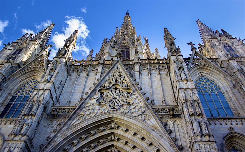 The richly decorated main facade of Barcelona's Catedral, dotted with spires, gargoyles and intricate stonework. Above the cathedral is a bright blue sky.