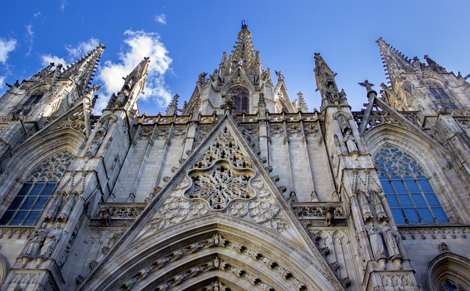 The richly decorated main facade of Barcelona's Catedral, dotted with spires, gargoyles and intricate stonework. Above the cathedral is a bright blue sky.
