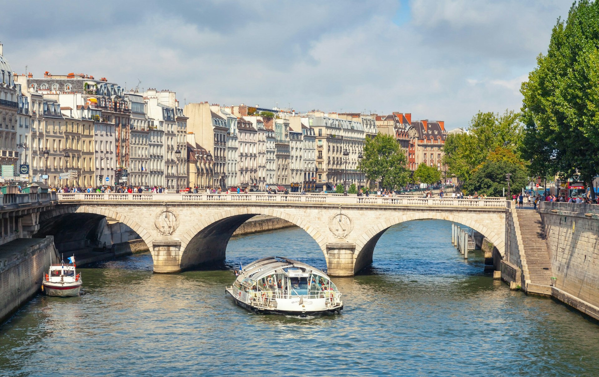 A Batobus tourist boat sailing under the stone arched Pont Neuf in Paris; on the left hand side of the river are handsome period buildings while the right hand side is lined with trees.