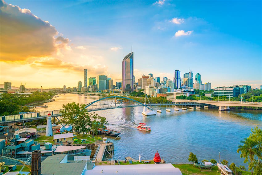 Looking down over the Brisbane River, with the city skyline of large glass towers in the background; the sky ranges from deep blue on the right to burning yellowy orange on the left, with the sun behind a bank of cloud; a small ferry makes its way under a bridge and towards the camera