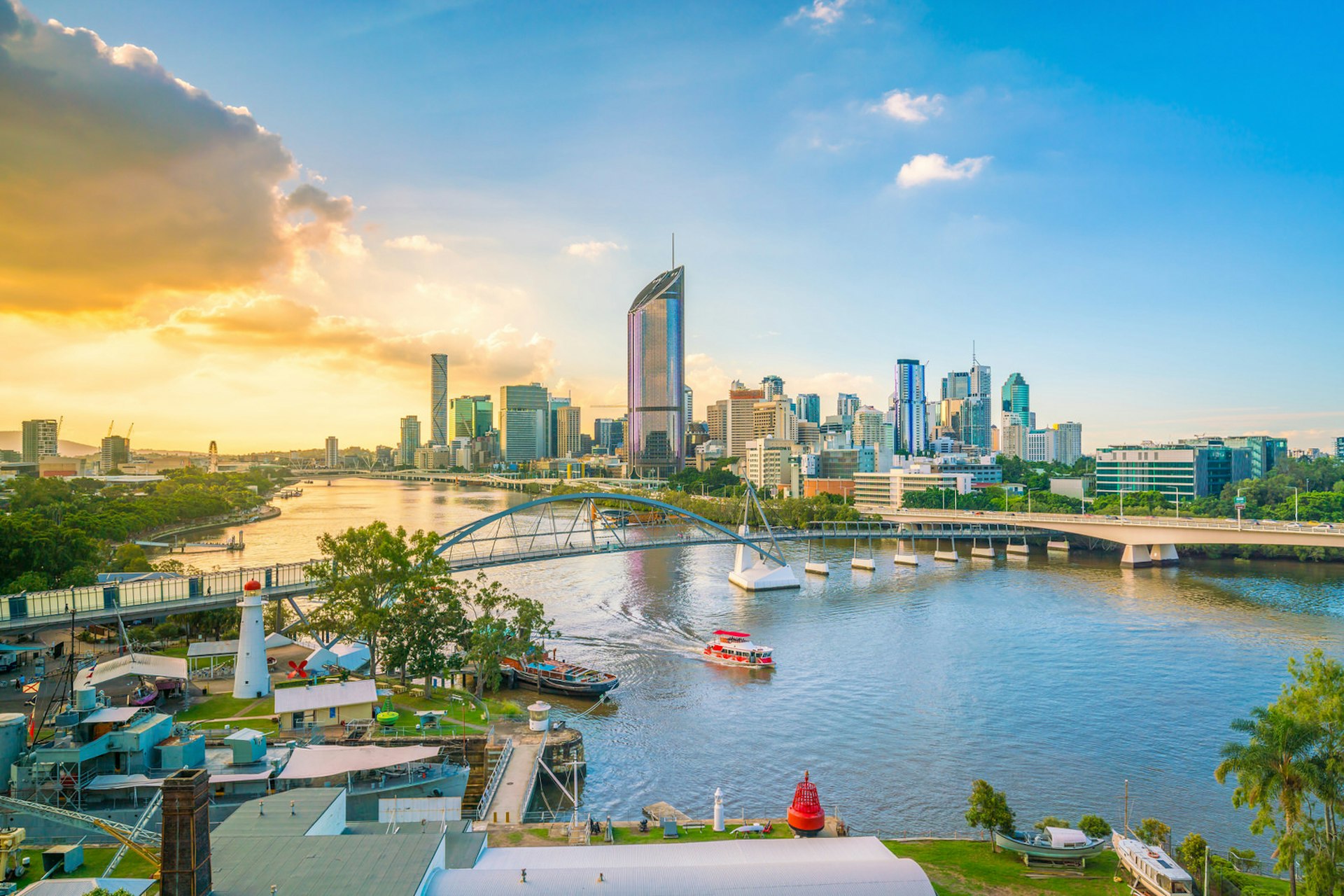 Looking down over the Brisbane River, with the city skyline of large glass towers in the background; the sky ranges from deep blue on the right to burning yellowy orange on the left, with the sun behind a bank of cloud; a small ferry makes its way under a bridge and towards the camera