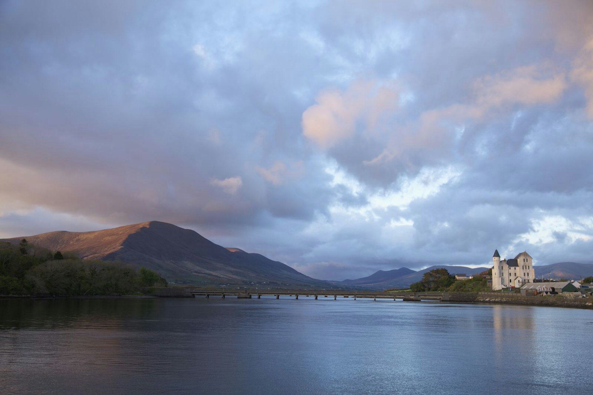 A museum made of white stone in on the right side of a river, joined by a bridge to the other side. In the background, a mountain looms over in the pink light of dusk.