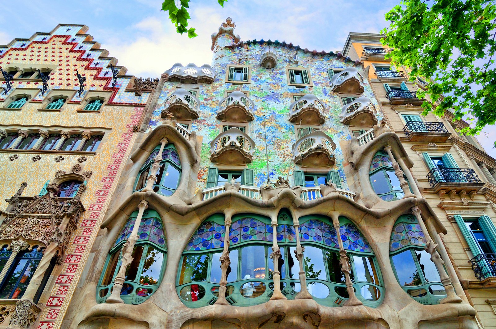 A low-level view of the whimsical facade of Casa Batlló in Barcelona; it is sprinkled with bits of blue, mauve and green tiles and studded with wave-shaped window frames and balconies, rising to an uneven roof with a solitary tower.