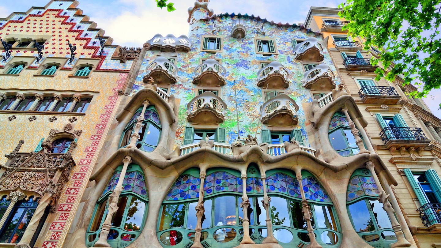 A low-level view of the whimsical facade of Casa Batlló in Barcelona; it is sprinkled with bits of blue, mauve and green tiles and studded with wave-shaped window frames and balconies, rising to an uneven roof with a solitary tower.