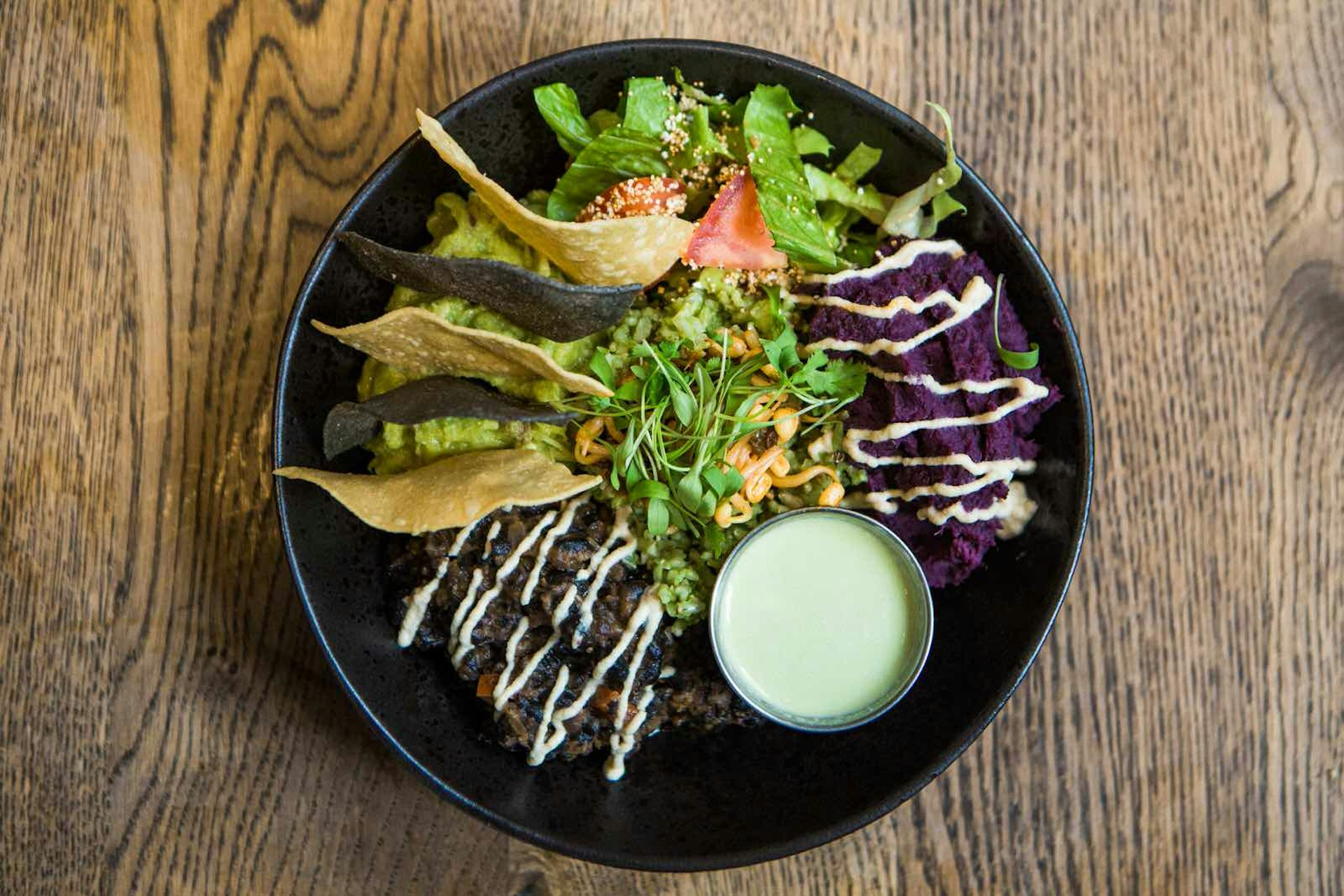 The Mexican bowl at Farmacy: sprouted coriander rice, romaine lettuce, tomatoes, guacamole, frijoles, chipotle ‘sour cream’, whipped squash, corn chips jalapeño dressing, arranged in a black bowl on a wooden table.