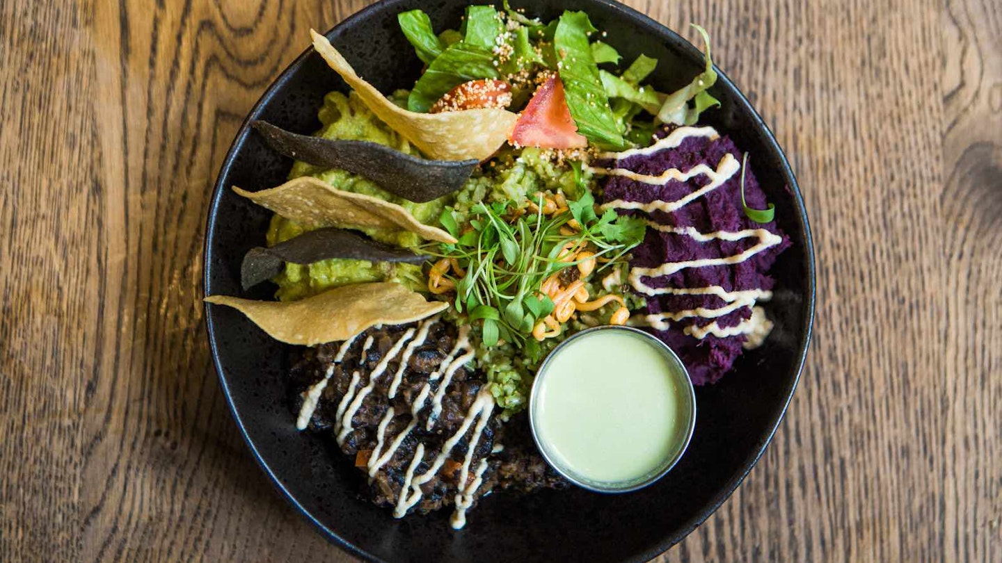 The Mexican bowl at Farmacy: sprouted coriander rice, romaine lettuce, tomatoes, guacamole, frijoles, chipotle ‘sour cream’, whipped squash, corn chips jalapeño dressing, arranged in a black bowl on a wooden table.