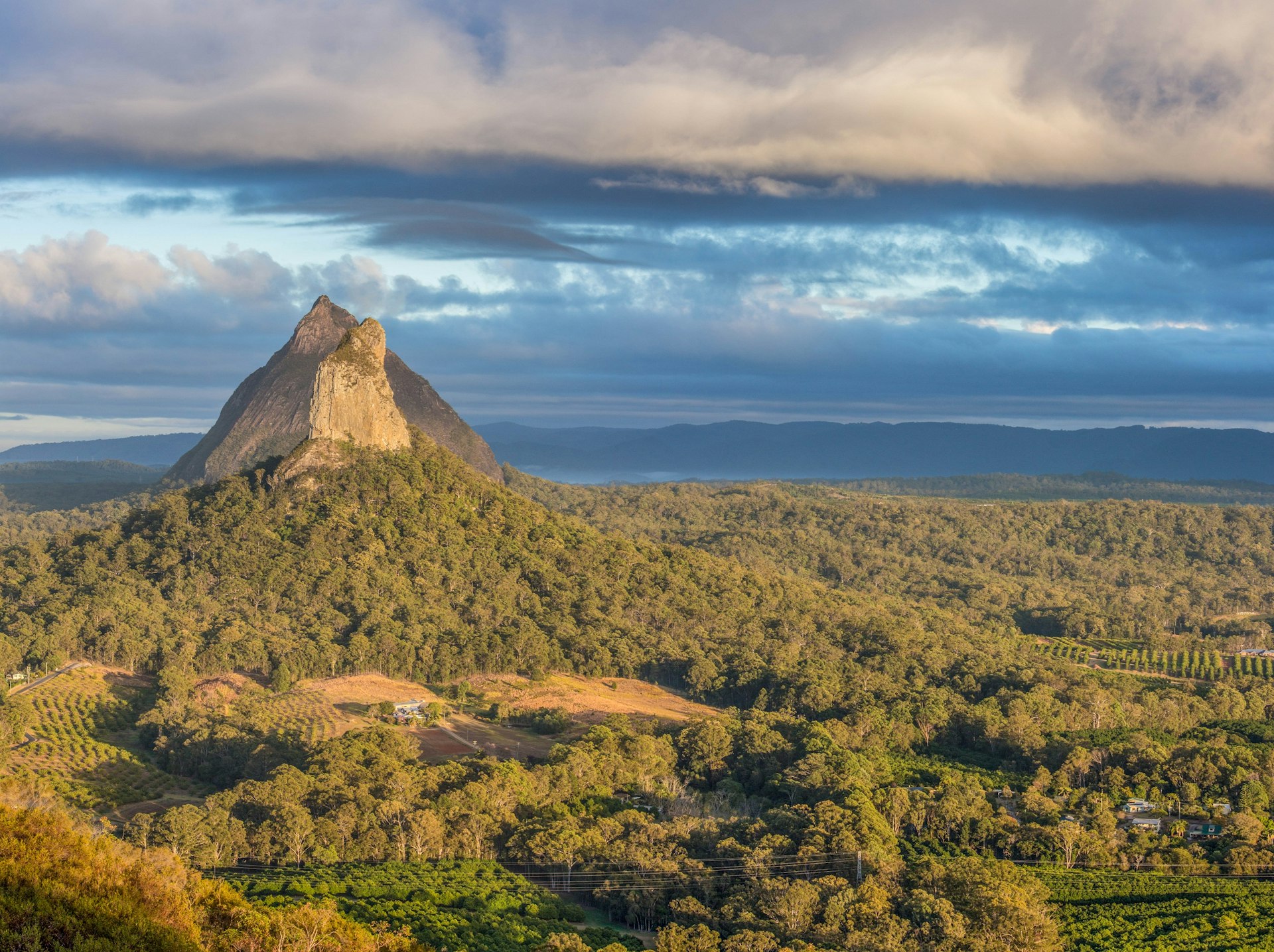 Rising pointedly out of a sea of forest and into a moody sky of dark clouds, pulled like cotton candy across a blue sky, is a rocky volcanic plug in Brisbane's Hinterland; like patches on a quilt, there are a few orchards within the foreground sections of forest