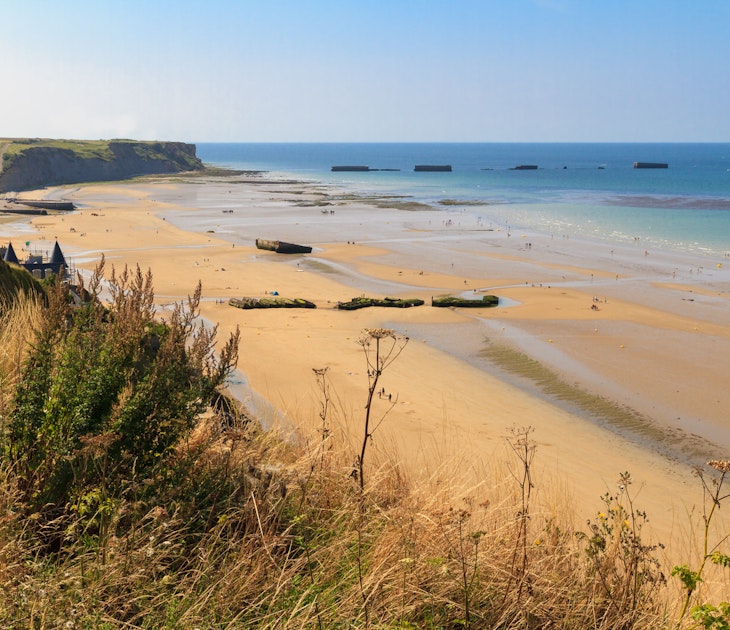 Looking down from a grassy clifftop over a huge golden D-Day landing beach, with the remains of a Mulberry Harbour (artificial port) at Arromanches-les-Bains visible in the distance