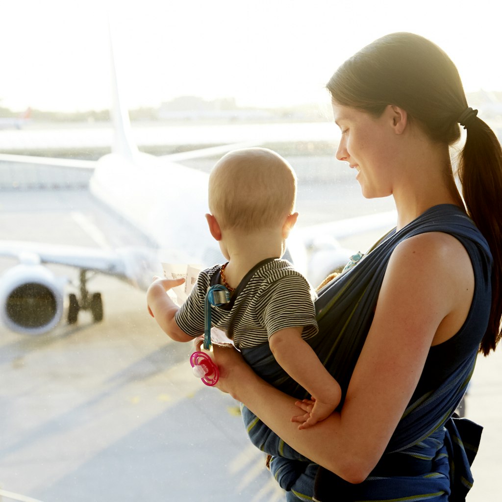 A mum looks lovingly down towards her young baby in a sling on her chest; the baby is looking away from the camera and out a window in the airport to a jet on the tarmac