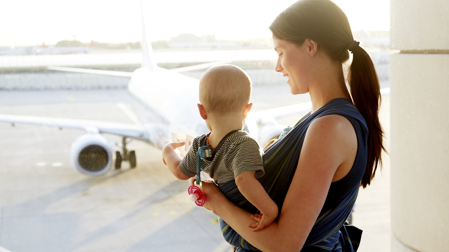A mum looks lovingly down towards her young baby in a sling on her chest; the baby is looking away from the camera and out a window in the airport to a jet on the tarmac