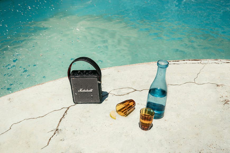 Produktbild av Stockwell II-högtalaren vid en pool bredvid en blåtonad karaff med vatten och två bruntonade koppar, varav en har vält;  Fars dag presenter