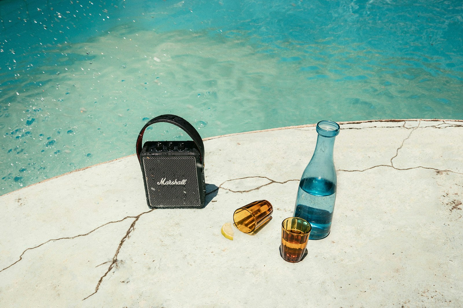 Product shot of the Stockwell II speaker by a pool next to a blue-tinted carafe of water and two brown-tinted cups, one of which has toppled over; Father's Day gifts