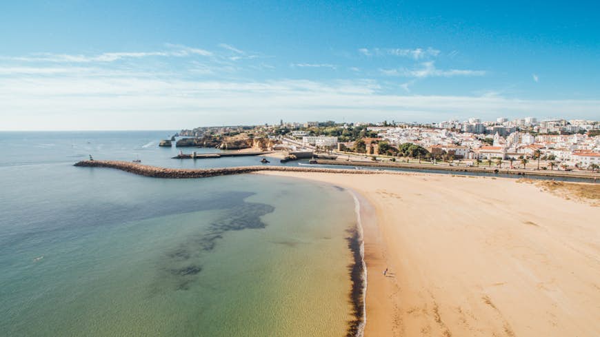 An aerial view of beautiful Meia Praia beach in the Algarve town of Lagos in the morning: a harbour wall and a river separate a huge deserted expanse of sand from the white buildings and ochre roofs of the town beyond, while a bright blue sky shines down on crystal-clear waters.