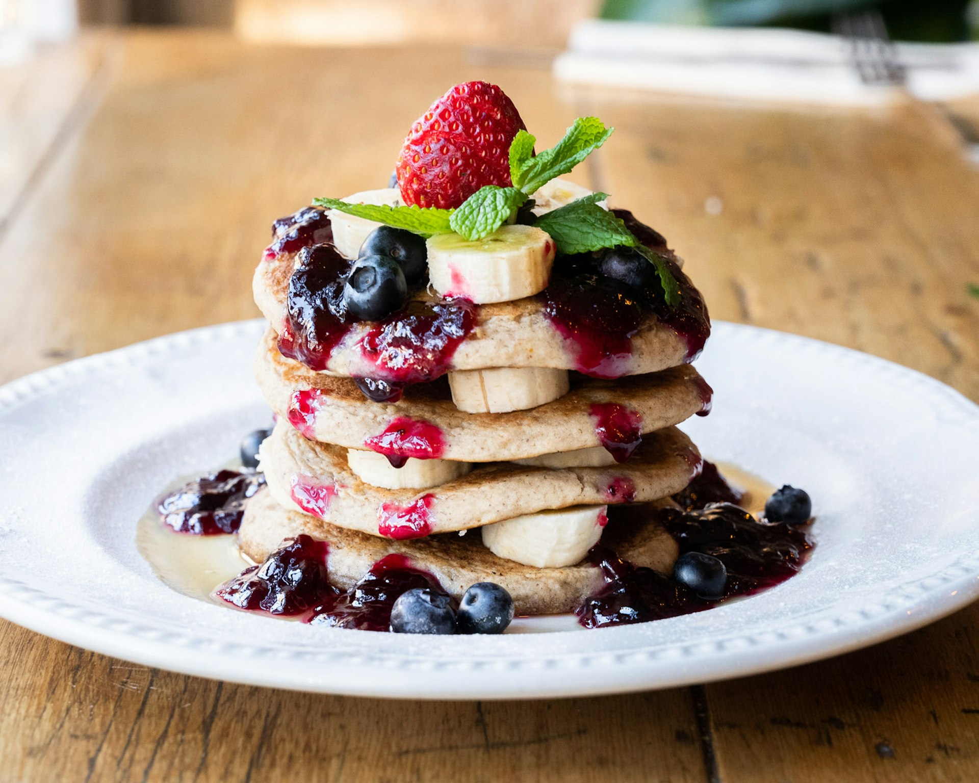 A huge stack of vegan pancakes at the Miranda Cafe, served with bananas, blueberries and a red compote, on a white plate dusted with icing sugar.