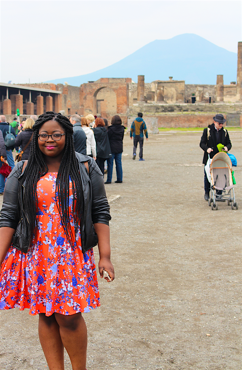 A young black woman poses in the foreground and some ruins in Pompeii are visible in the background. Other visitors mingle around; she is standing alone. Mount Vesuvius looms in the background of the photo. A black girl travelling through parts of Italy may experience racism. 