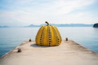 A giant yellow and black pumpkin sculpture at the end of a concrete jetty in front of the sea in Naoshima, with hilly islands visible in the background