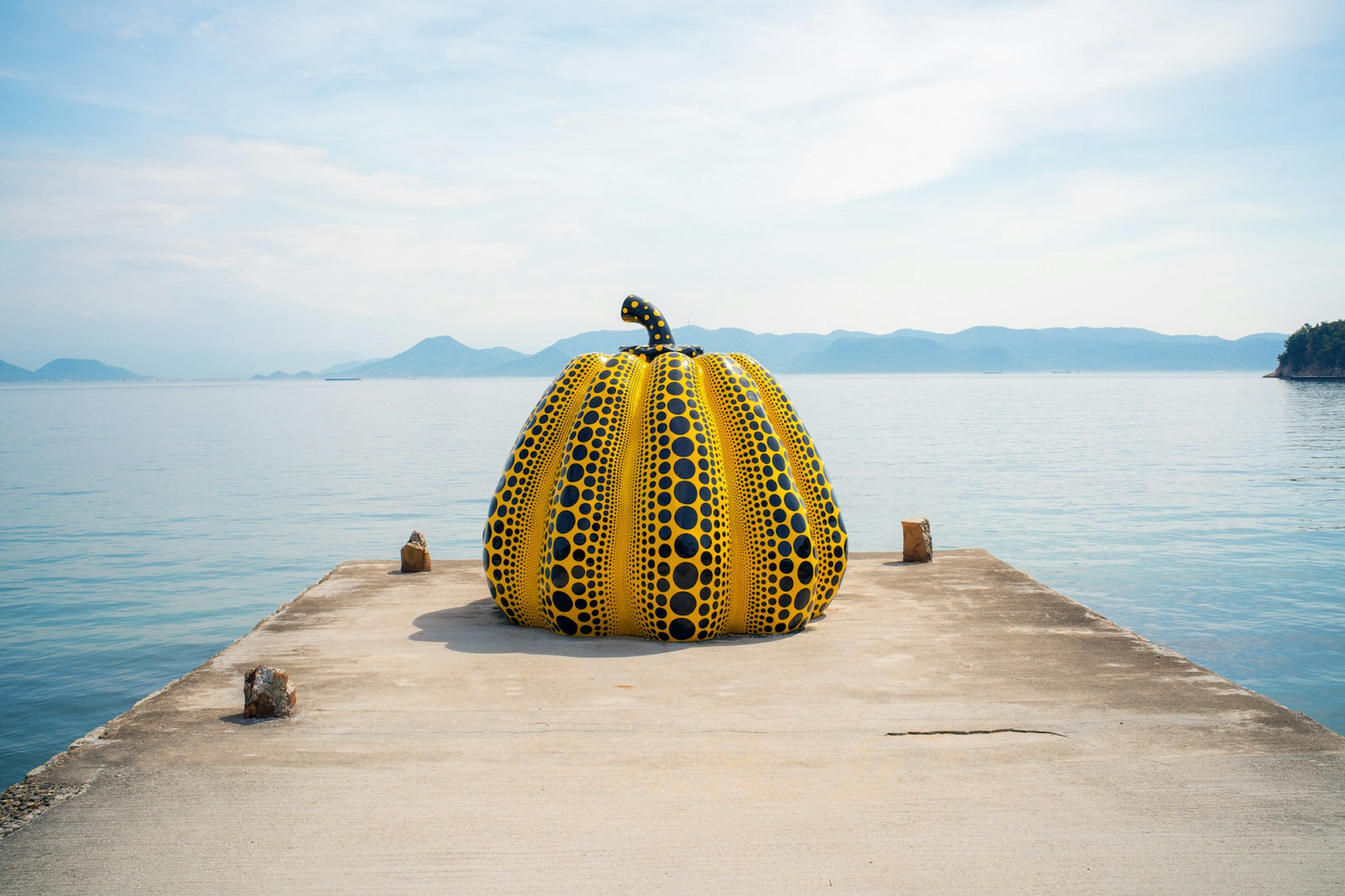 A giant yellow and black pumpkin sculpture by Yayoi Kusama at the end of a concrete jetty in front of the sea in Naoshima, with hilly islands visible in the background