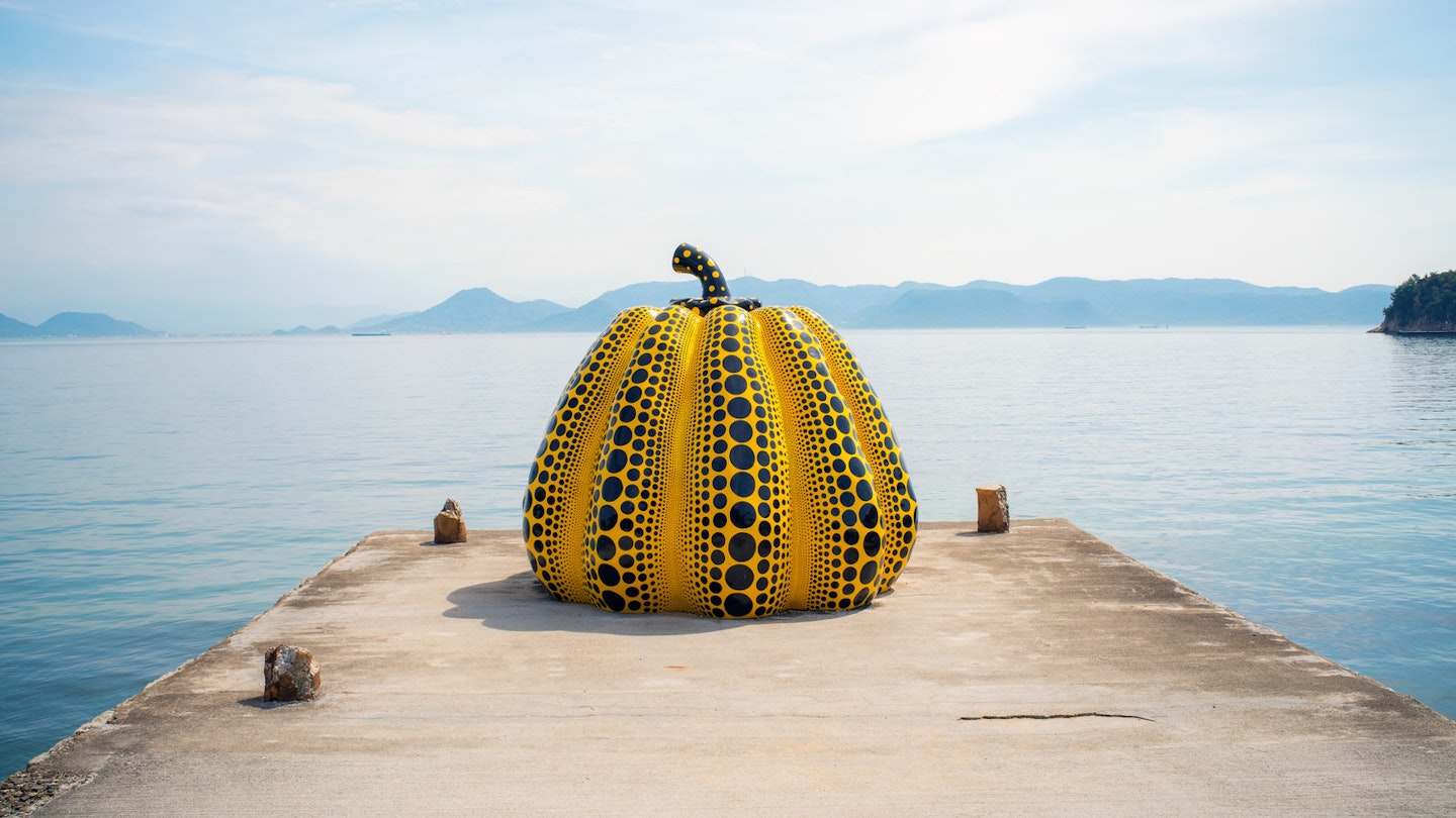 A giant yellow and black pumpkin sculpture at the end of a concrete jetty in front of the sea in Naoshima, with hilly islands visible in the background