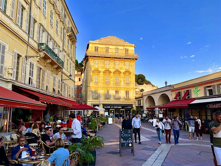 A pedestrian street in Old Nice at sunset. There are lots of outside tables for restaurants filled with people and others are walking down the street with the light hitting off a building in the background.