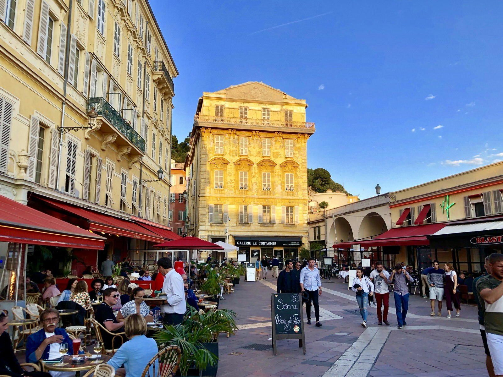 A pedestrian street in Old Nice at sunset. There are lots of outside tables for restaurants filled with people and others are walking down the street with the light hitting off a building in the background.