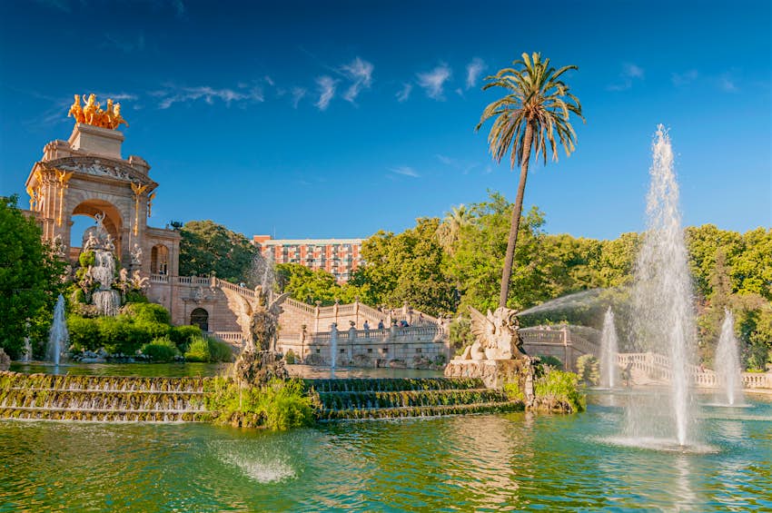 A perfect weekend in Barcelona should take in the monumental waterfall at Parc de la Ciutadella, a dramatic combination of statuary, rugged rocks, greenery and water; there are trees and a modern building in the background and the greenish water of a lake in the foreground.