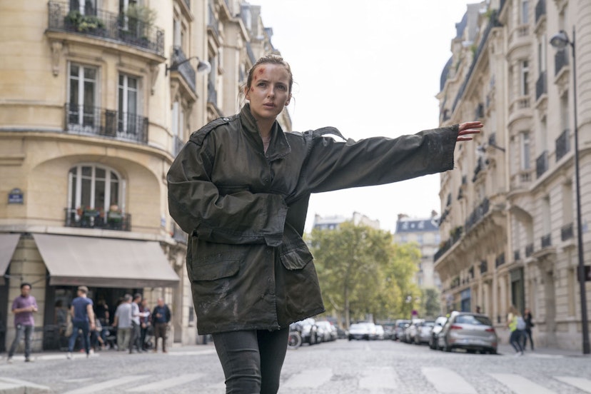 A still from the opening episode of Killing Eve season two: Villanelle (played by Jodie Comer) stands on a Paris street wearing a green coat; she has a head injury and one arm outstretched as if to flag down a taxi.