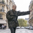 A still from the opening episode of Killing Eve season two: Villanelle (played by Jodie Comer) stands on a Paris street wearing a green coat; she has a head injury and one arm outstretched as if to flag down a taxi.