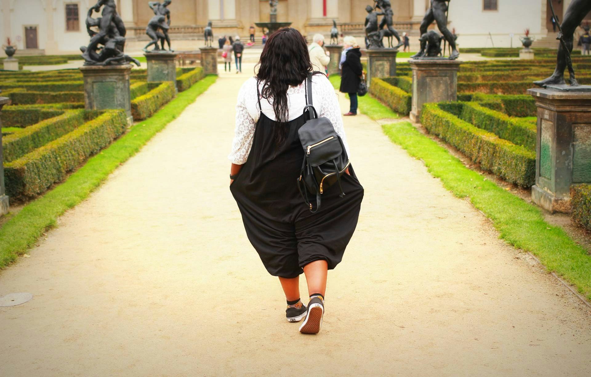 A black girl travelling through Prague. She's wearing a backpack walks along a path in a public garden in Prague. She is facing away from the camera.