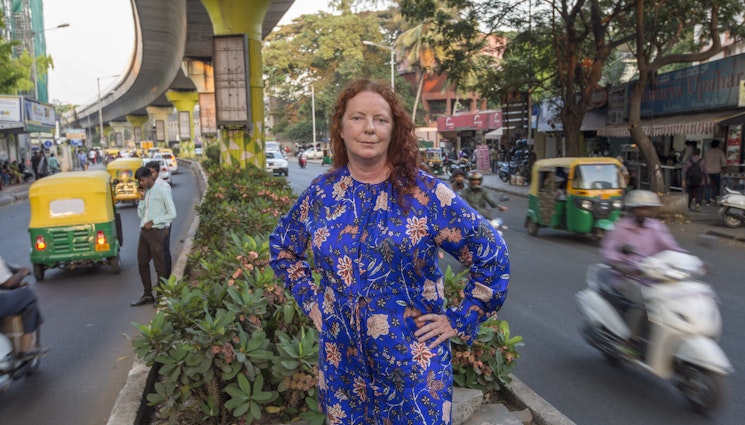 The author poses in India. She is standing in an island in the middle of a road, wearing a blue dress. Tuk tuks, mopeds and cars are speeding past her on either side.