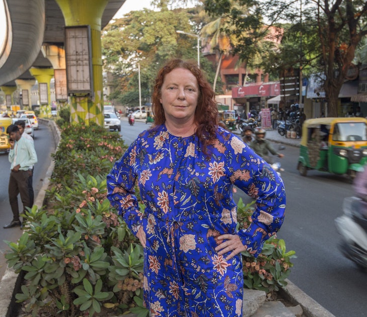 The author poses in India. She is standing in an island in the middle of a road, wearing a blue dress. Tuk tuks, mopeds and cars are speeding past her on either side.