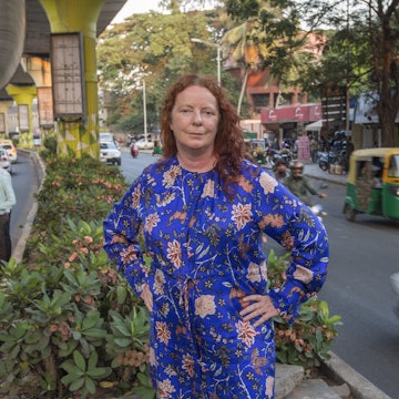 The author poses in India. She is standing in an island in the middle of a road, wearing a blue dress. Tuk tuks, mopeds and cars are speeding past her on either side.