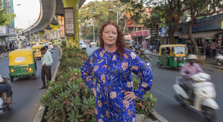 The author poses in India. She is standing in an island in the middle of a road, wearing a blue dress. Tuk tuks, mopeds and cars are speeding past her on either side.