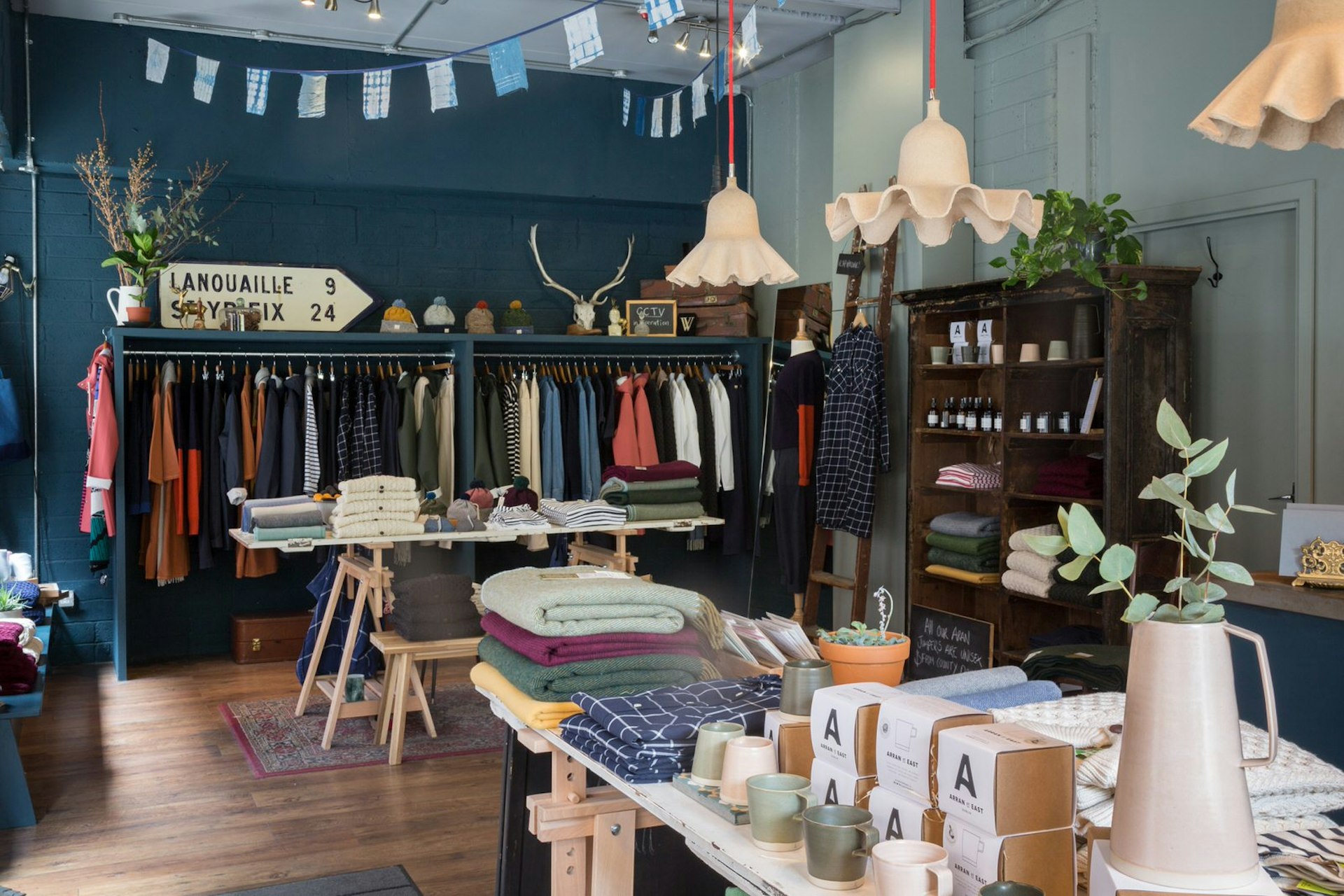 Dublin independent shops - the interior of Scout. There is a rail of multicoloured clothes against a teal wall with a road sign and horned skull on top. Blue bunting hangs from the roof and there is a table of folded clothes, mugs and homewares in the foreground as well as a variety of lampshades hanging at different lengths from the roof