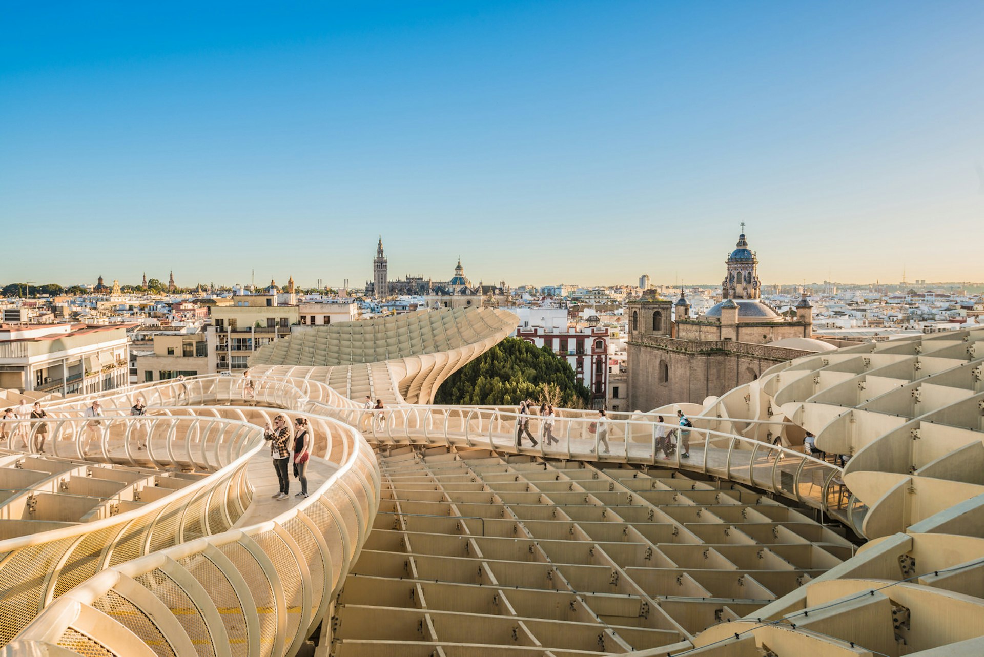 People walking on top of the Metropol Parasol, a huge structure made of wood and concrete in the shape of a pergola; there is a beautiful view across the Seville skyline, taking in the cathedral and Giralda bell tower.