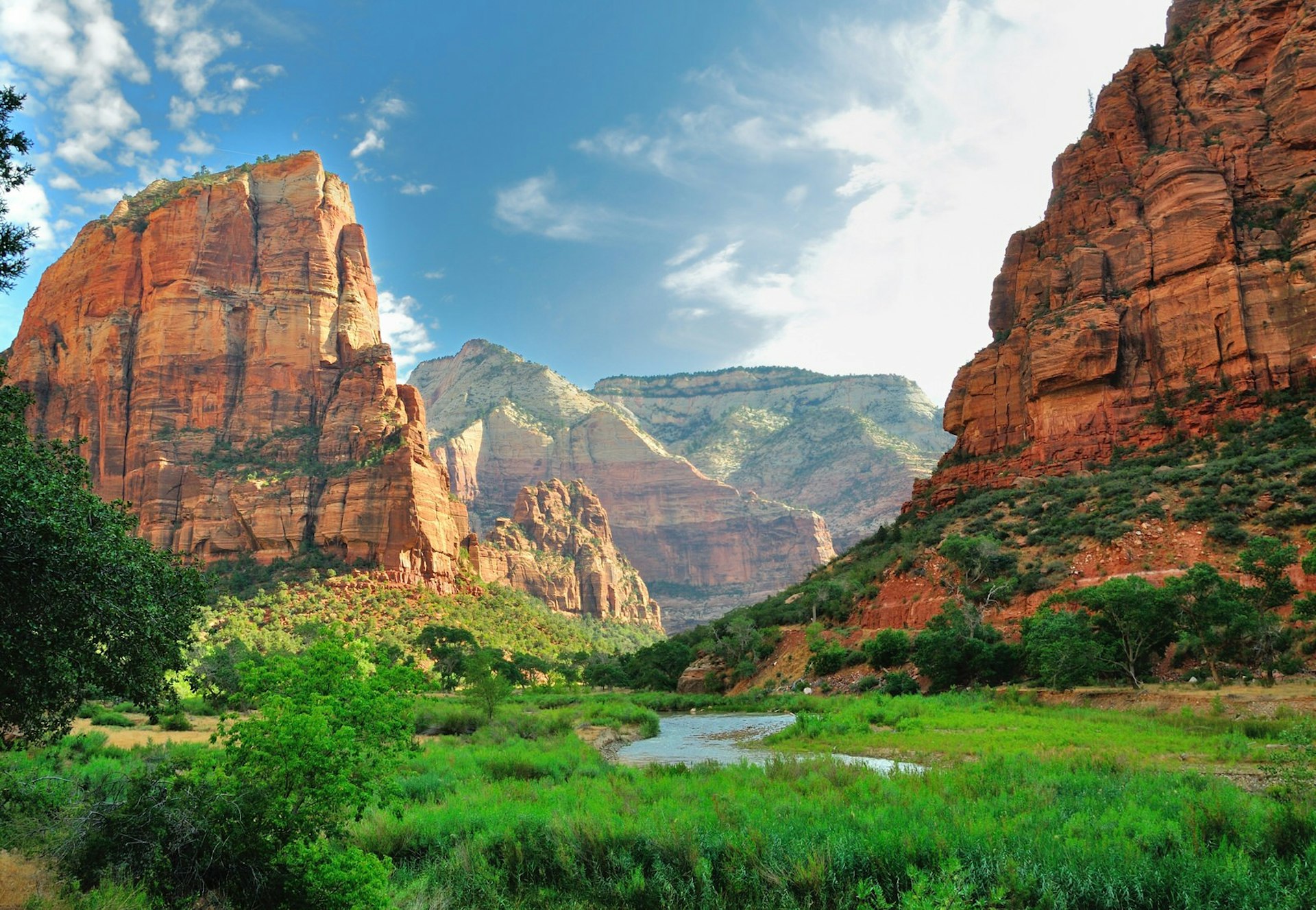 In Utah's Zion National Park, huge ochre cliffs mottled with greenery rise from a verdant valley floor, through which a narrow river bends; more cliffs are visible in the background under a blue sky.