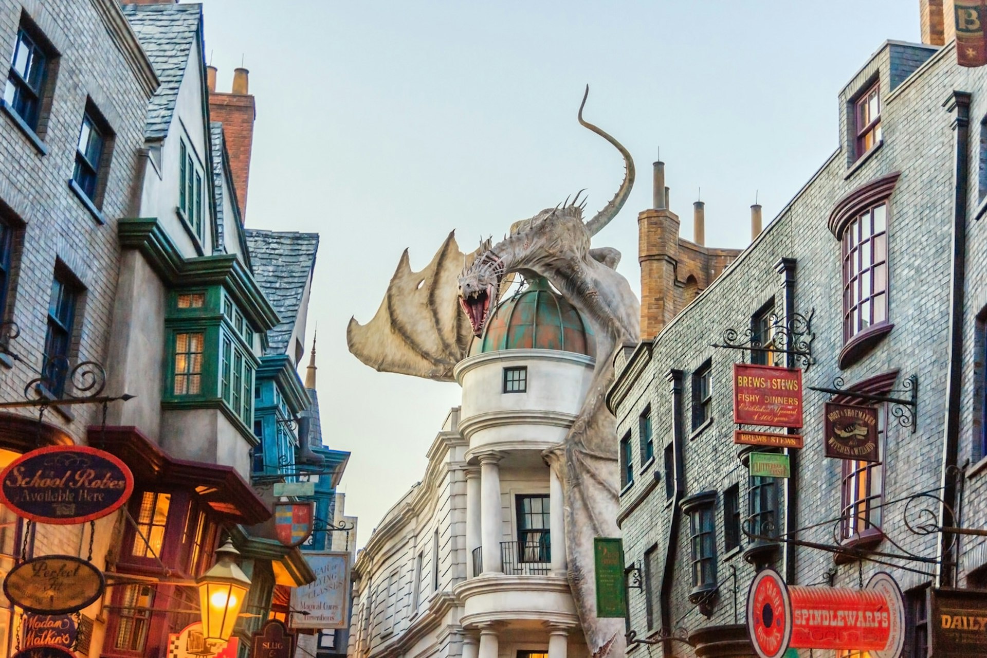 A street at the Wizarding World of Harry Potter in Florida, full of quaint, crooked olde-worlde houses and shops with signs hanging outside; the most notable building has a cupola on its roof which is being straddled by an angry-looking dragon.