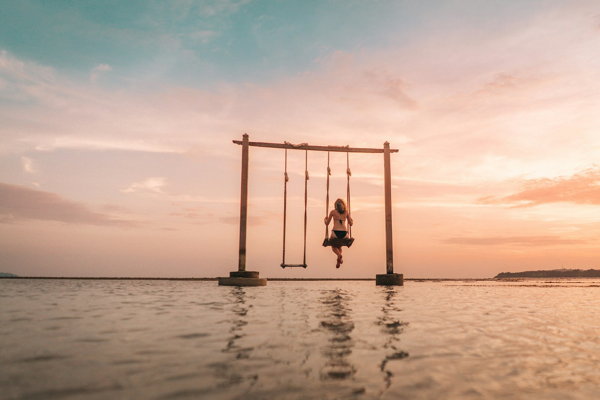 A beautiful pink sunset in Gili Air, Indonesia. There is a large double swing in the sea with the silhouette of a person on one of the swings.