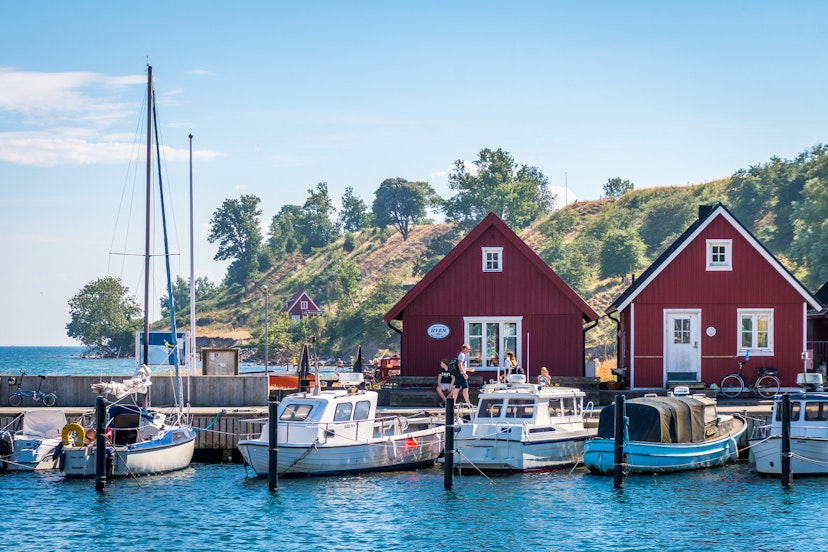 Small boats are docked in front of two red triangular buildings on a sunny day on Ven island, Sweden