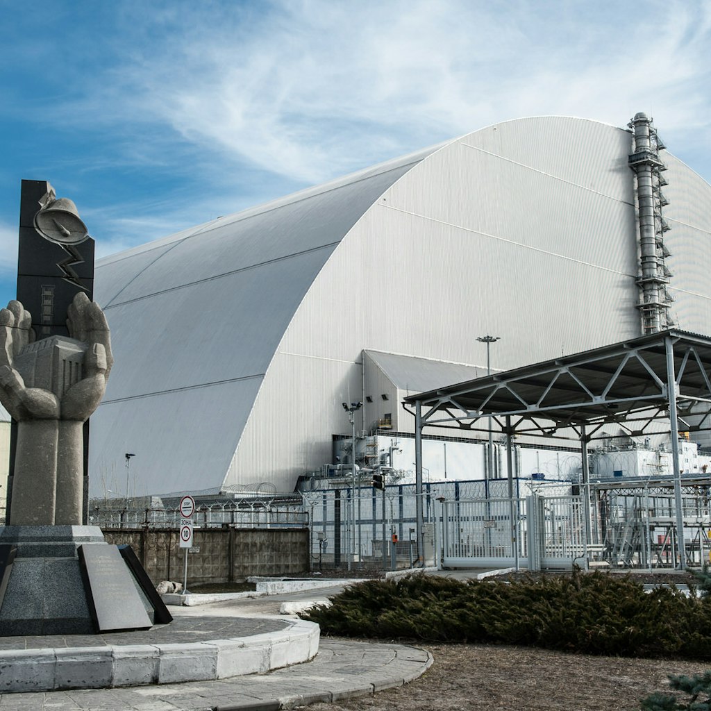 In the foreground is a memorial to the Chernobyl disaster, depicting two hands cupped together around a miniature of Reactor 4; behind the memorial is the huge grey arch of the New Safe Confinement structure which encloses the remains of Reactor 4.
