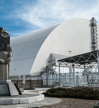 In the foreground is a memorial to the Chernobyl disaster, depicting two hands cupped together around a miniature of Reactor 4; behind the memorial is the huge grey arch of the New Safe Confinement structure which encloses the remains of Reactor 4.