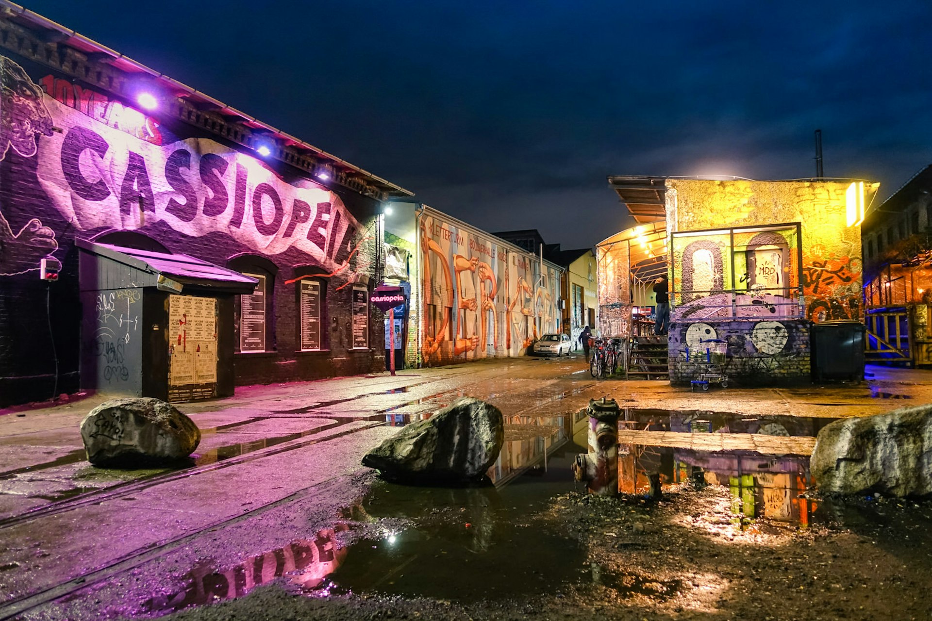 Berlin clubs - the main yard at RAW Gelände at night time. The buildings are illuminated by multicoloured lights and the club Cassiopeia has a huge mural of its name along the side of the building