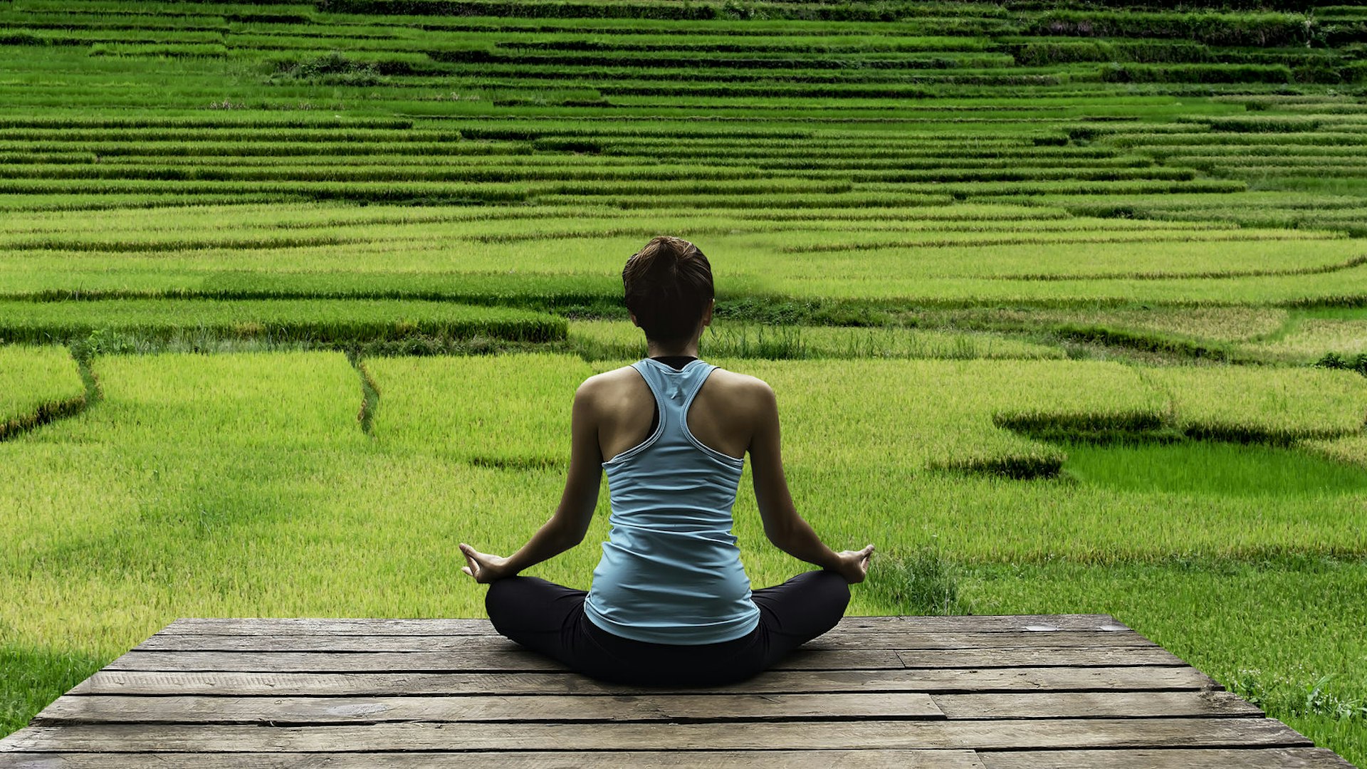 A woman, sitting on wooden decking, meditates while facing out towards rice fields in Bali
