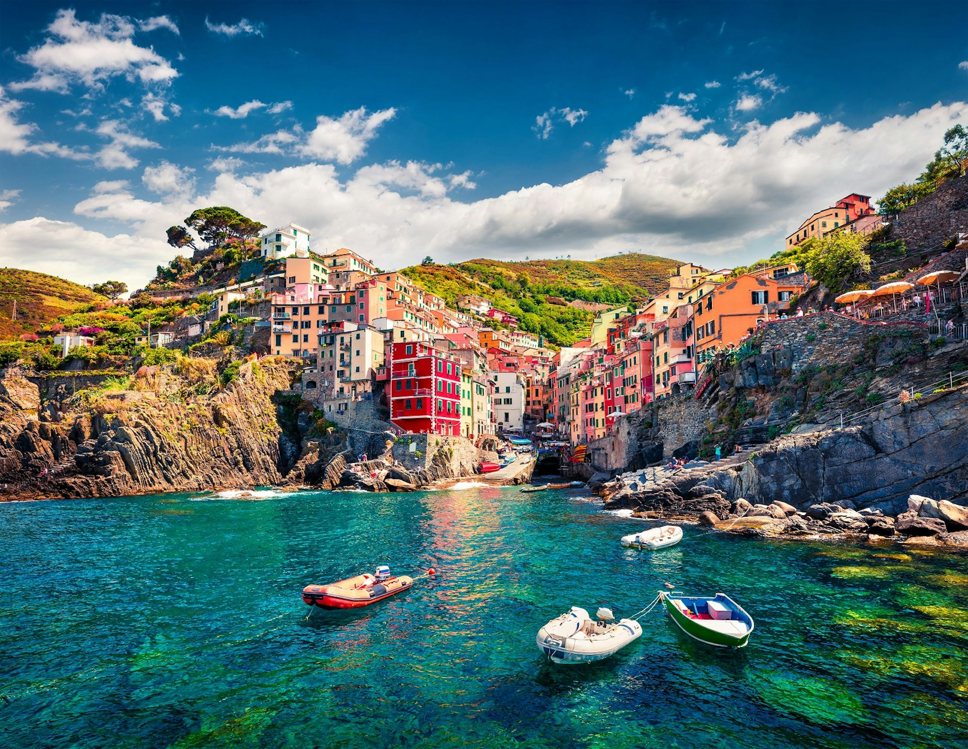 A view of the Cinque Terre, Italy, from the sea. There are a few dinghies moored in the foreground and beyond the beautiful blue water of the Mediterranean we can see the colourful hill cities