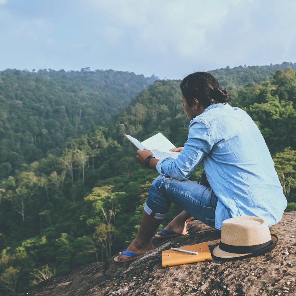 A person sitting on a rock reading a book next to a notepad, pen and hat. The person is wearing a denim shirt, jeans and flip flops and beyond the rock there is a view of rolling greenery