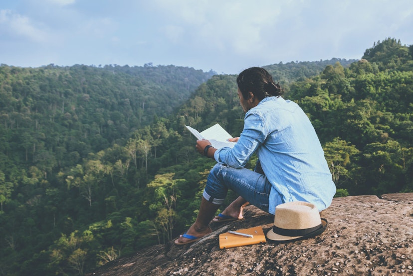 A person sitting on a rock reading a book next to a notepad, pen and hat. The person is wearing a denim shirt, jeans and flip flops and beyond the rock there is a view of rolling greenery