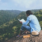 A person sitting on a rock reading a book next to a notepad, pen and hat. The person is wearing a denim shirt, jeans and flip flops and beyond the rock there is a view of rolling greenery