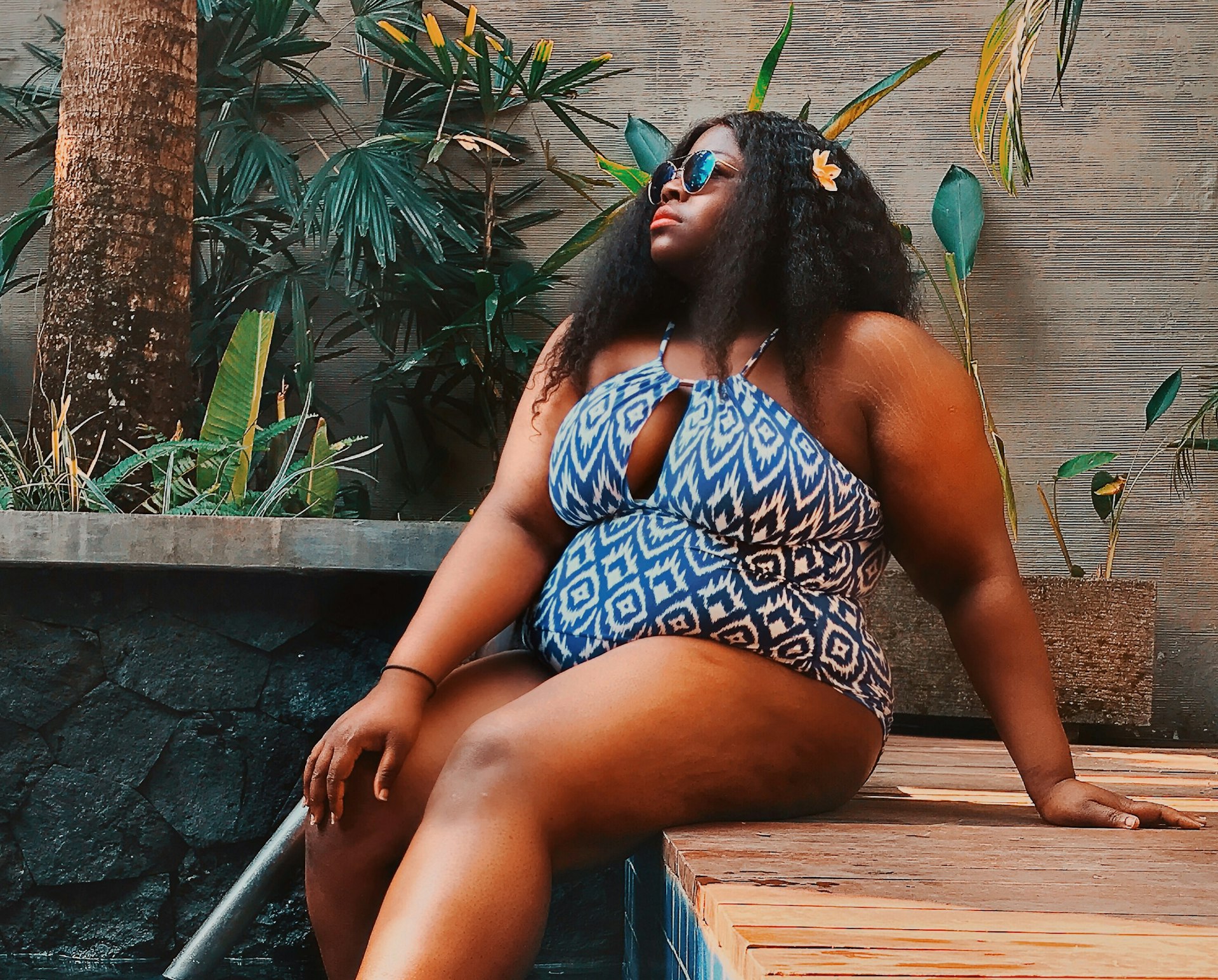 Writer Stephanie Yeboah relaxes by a pool in a blue-and-white patterned swimsuit and blue-lens sunglasses. There are palm-like trees in the background and she is sitting on a wooden deck with her feet dangling in the water