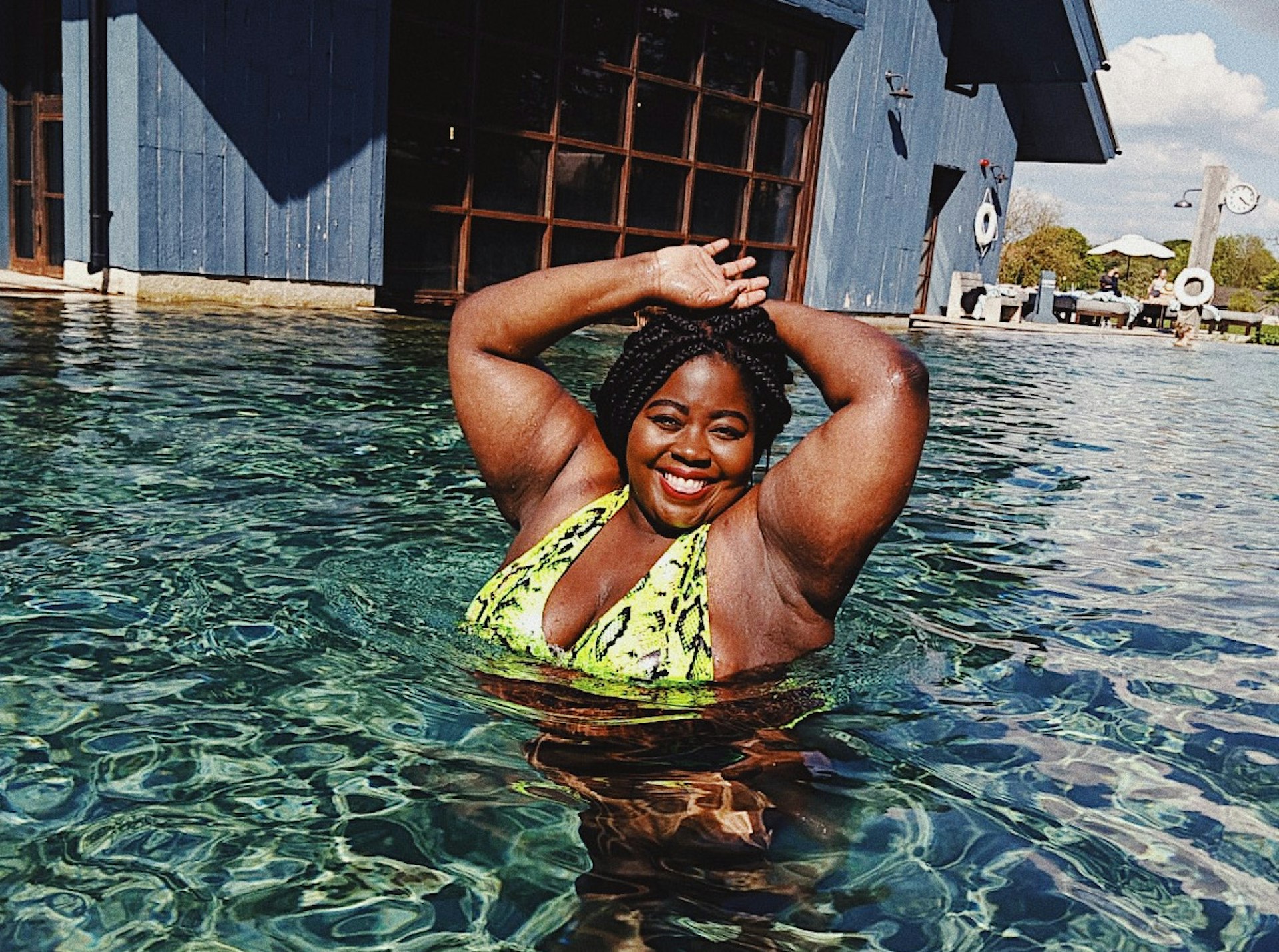 Stephanie Yeboah in a sun-drenched pool in a lime green snakeskin-print bikini. Stepahnie is smiling at the camera with her arms above her head and there is a blue building in the background