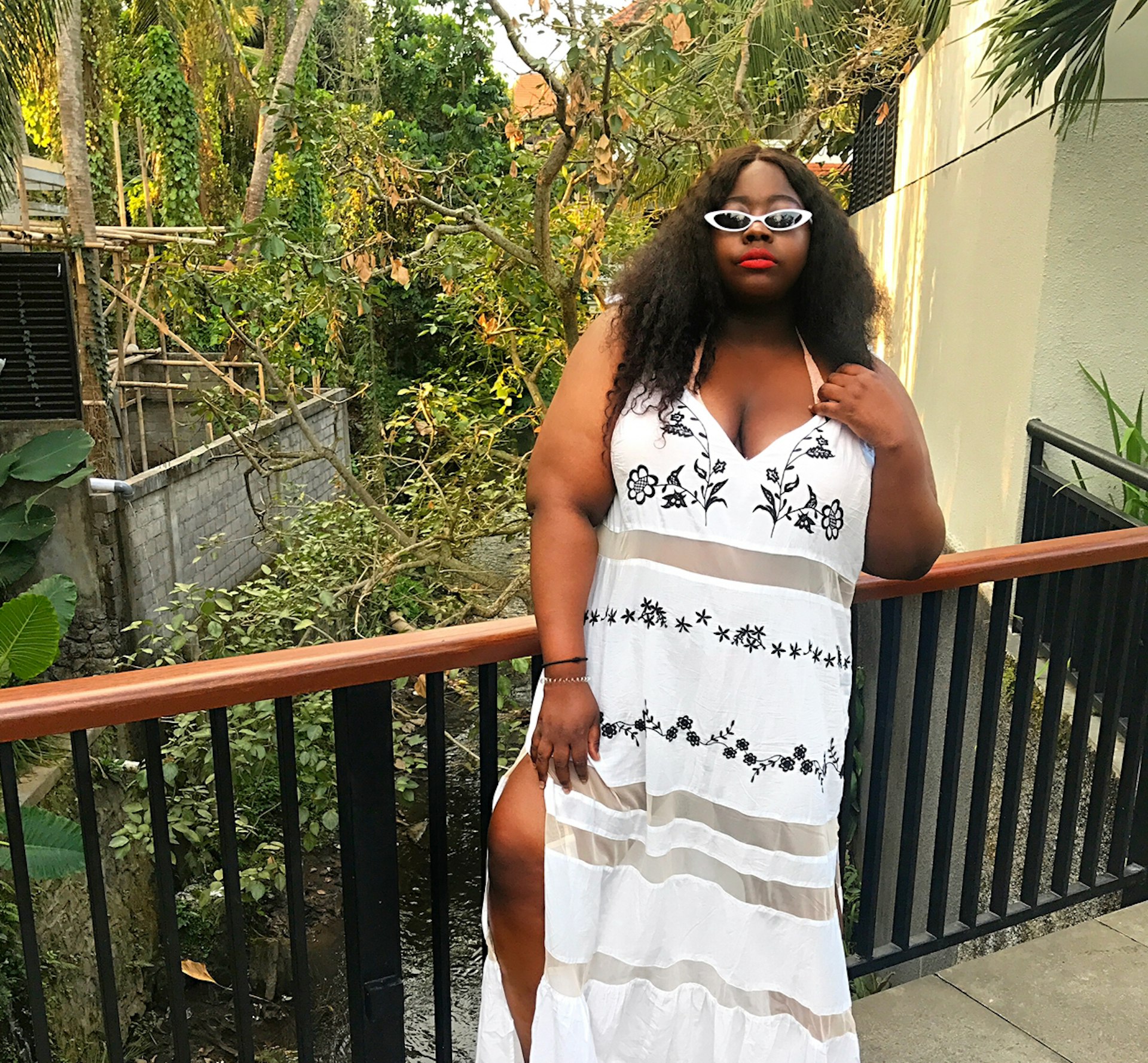 Stephanie Yeboah in a white sun dress with sheer vertical panels and black flower embroidery. She is wearing white-framed sunglasses and standing on a bridge in front of a small stream and greenery